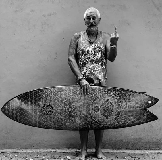 Old man holding a surfboard and flipping off the camera.