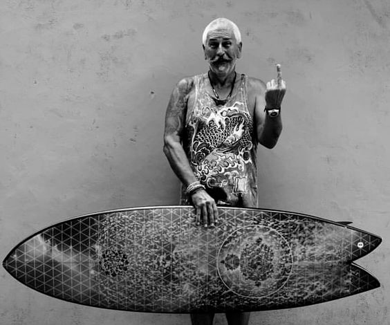 Old man holding a surfboard and flipping off the camera.